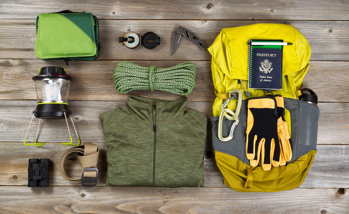 essential camping gear spread out on a wooden floor including backpack, clothing, lantern, ropes.