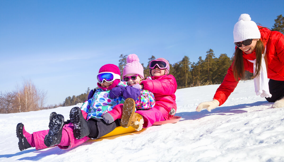 3 kids on a sled as their mother pushes them down a snowy hill