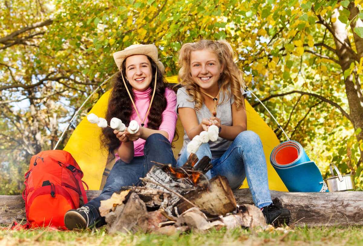 two teenage girls sitting in front of a yellow tent roasting marshmallows over a fire