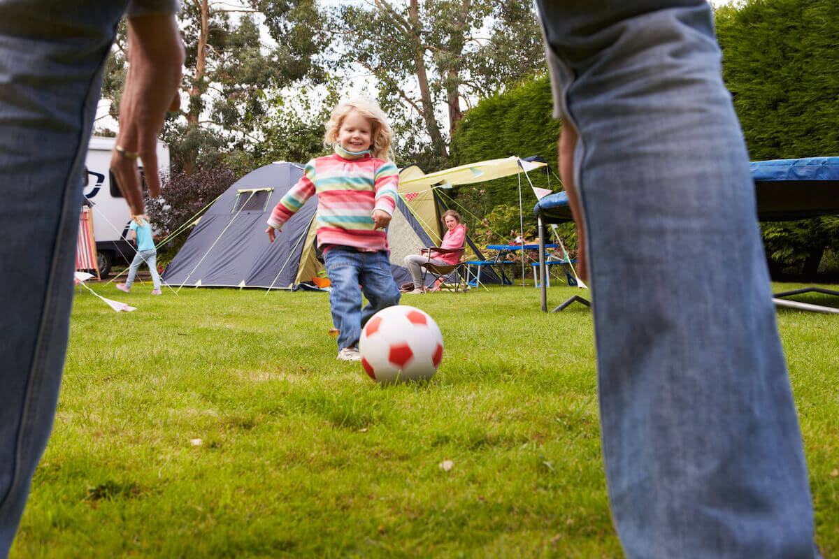 young child kicking a soccer ball to an adult at a camping site