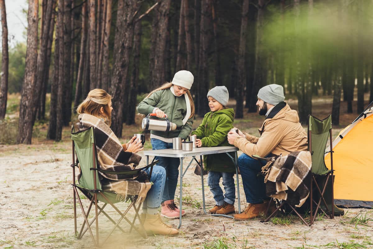 family sitting at table and chairs next to tent, pouring hot drink from a flask.