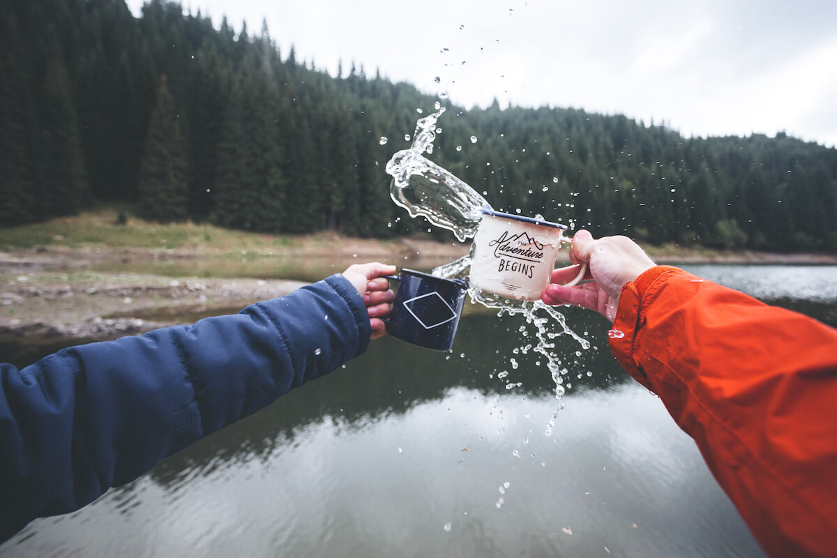 two people standing in front of a lake, knocking their camping mugs together.