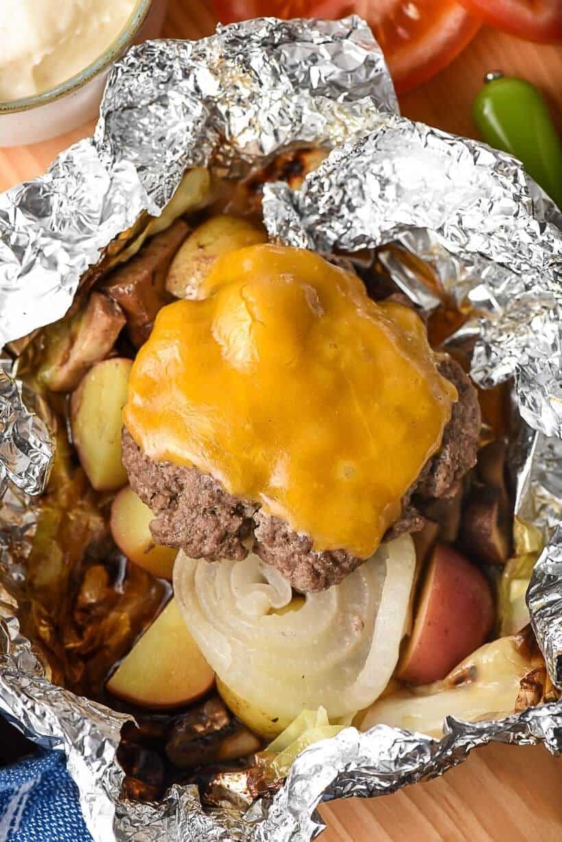 hamburger patty, with melted cheese slice on top sitting on top of roasted vegetables in foil.