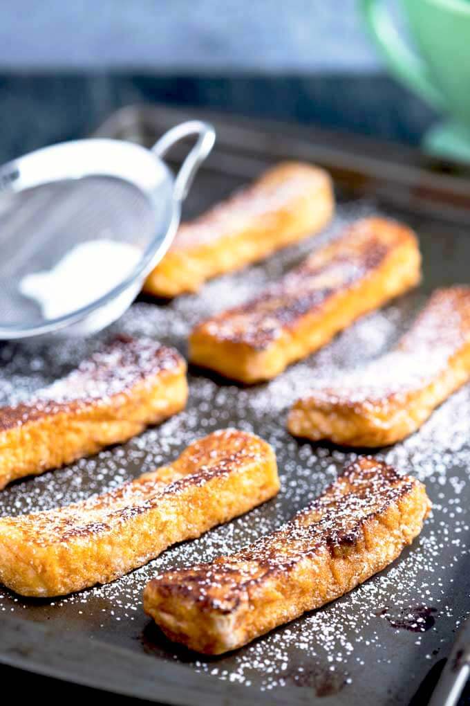 french toast fingers on a serving tray with icing sugar on top and a sifter in the background.