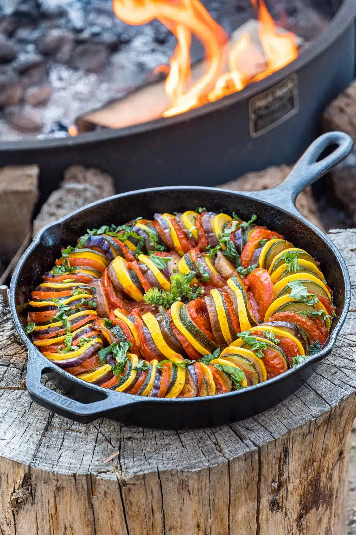 skillet of sliced vegetables on wooden table in front of campfire.