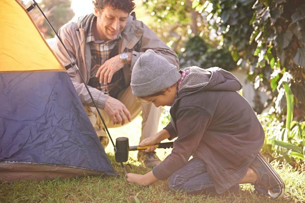 young boy in warm clothing learning how to hammer tent pegs in with his dad helping.