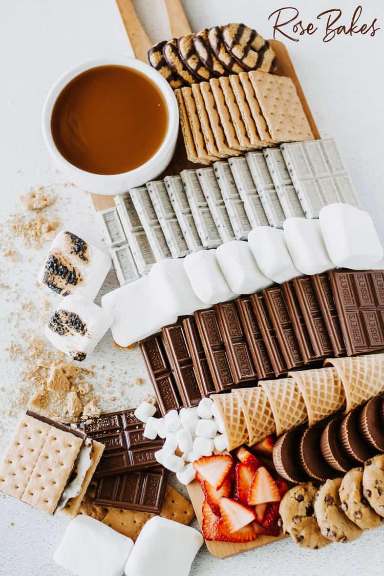 S'mores charcuterie board with chocolates, marshmallows, crackers and ice cream cones on a white board.
