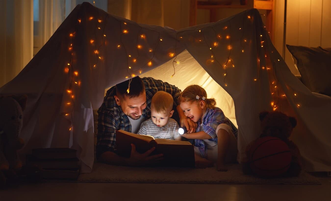 tent set up indoors with fairy lights and dad reading to kids inside.